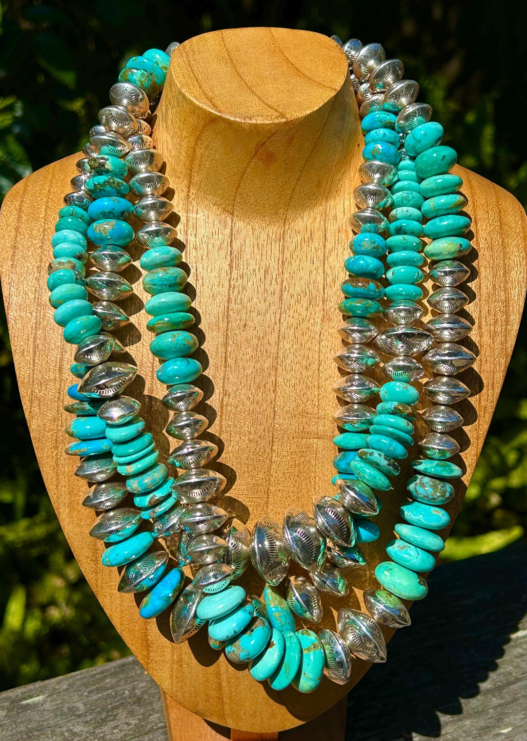 May include: A turquoise and silver necklace with multiple strands of oval-shaped turquoise beads and silver beads with a stamped design. The necklace is displayed on a wooden mannequin.