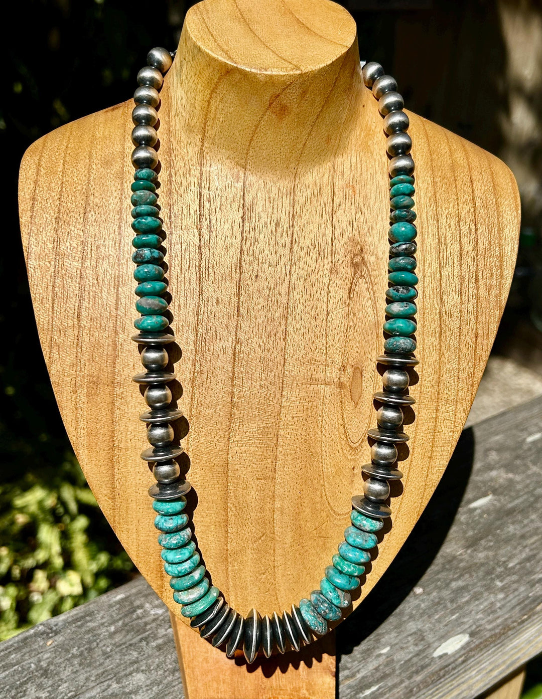 May include: A turquoise and silver necklace with round beads and flat, round turquoise stones. The necklace is displayed on a wooden mannequin.