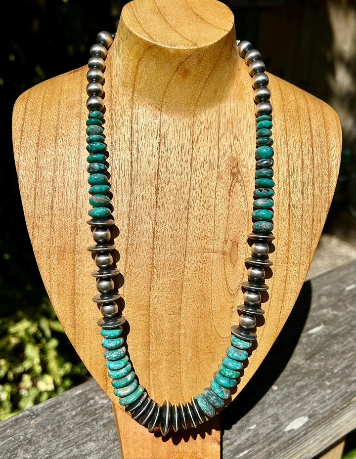 May include: A turquoise and silver necklace with round beads and flat, round turquoise stones. The necklace is displayed on a wooden mannequin.