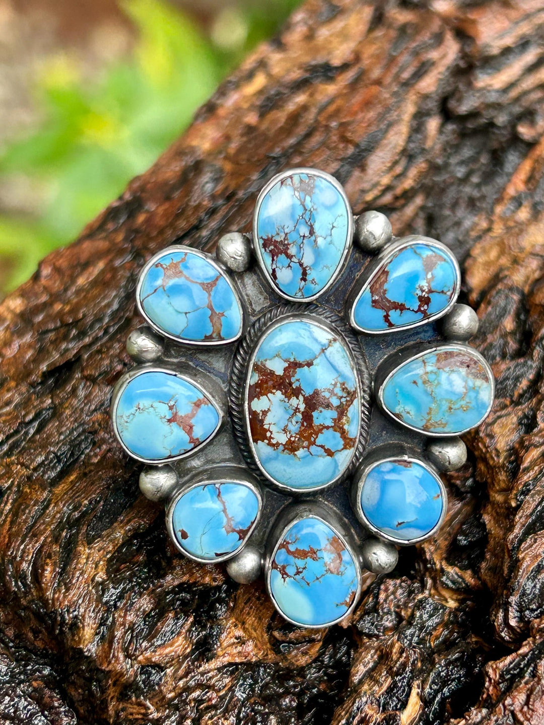 May include: A silver ring with a cluster of nine turquoise stones. The stones are a light blue color with brown veins and are set in a silver bezel setting. The ring is on a brown wooden surface.
