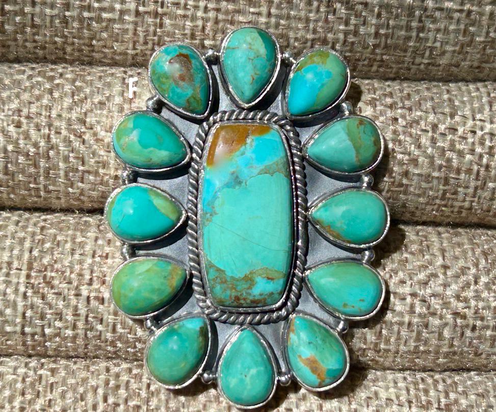 May include: A silver ring with a large, oval turquoise stone in the center, surrounded by smaller turquoise stones. The central stone is a vibrant teal color with brown marbling, while the surrounding stones are a lighter shade of teal. The ring has a detailed, ornate design.